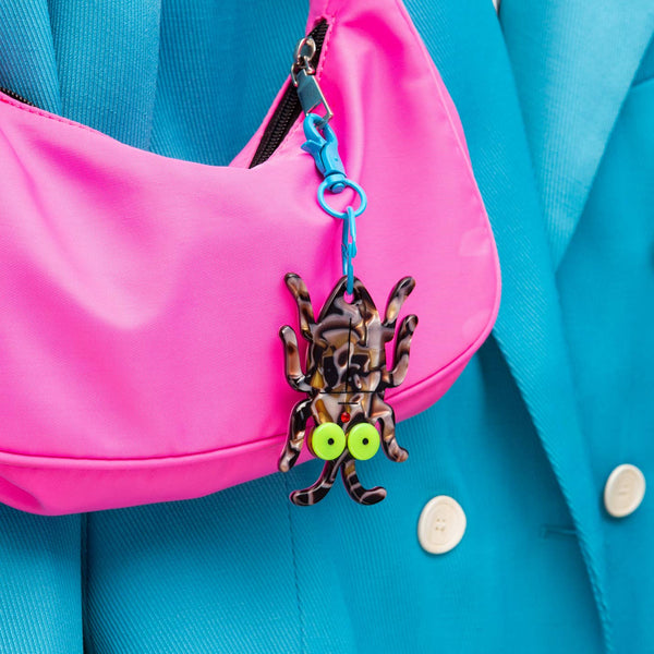 Acrylic swirled brown and black cockroach-shaped keychain with large lime green eyes and red rhinestone mouth on a bright blue enameled metal swivel keychain. Shown on a pink purse