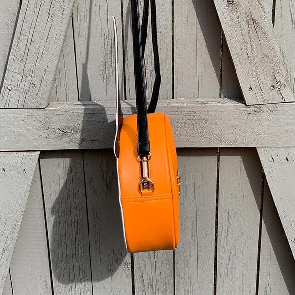 Faux leather shoulder bag in the shape of an orange jack-o’-lantern pumpkin with smiling face and black witch hat with yellow stars. Bag has adjustable black faux leather strap. Shown from side hanging on fence to display strap hardware and exterior zipper 