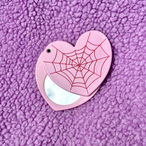 Light pink heart-shaped compact pocket mirror with red spider web design on a purple textured background