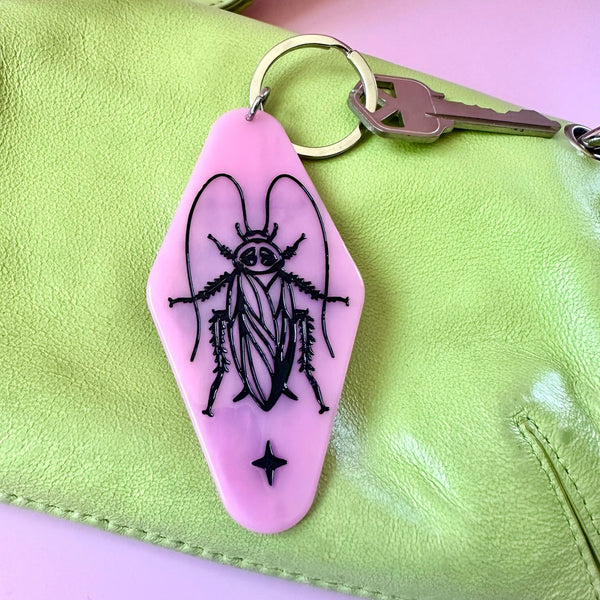 A pink hotel-style keychain featuring a black etched illustration of a cockroach, shown attached to a key ring on a light green leather bag against a pink background.