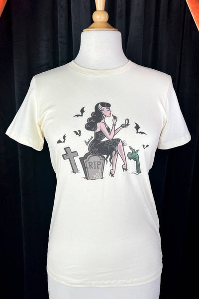 off-white fitted t-shirt with illustration of a woman sitting on a gravestone in a cemetery wearing a spiderweb patterned wiggle dress holding a compact mirror and applying lipstick surrounded by bats and a zombie hand. Shown on dress form 