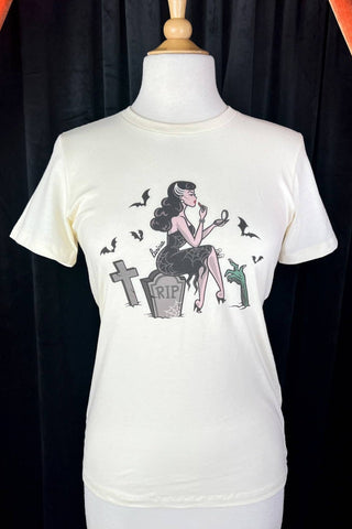 off-white fitted t-shirt with illustration of a woman sitting on a gravestone in a cemetery wearing a spiderweb patterned wiggle dress holding a compact mirror and applying lipstick surrounded by bats and a zombie hand. Shown on dress form 