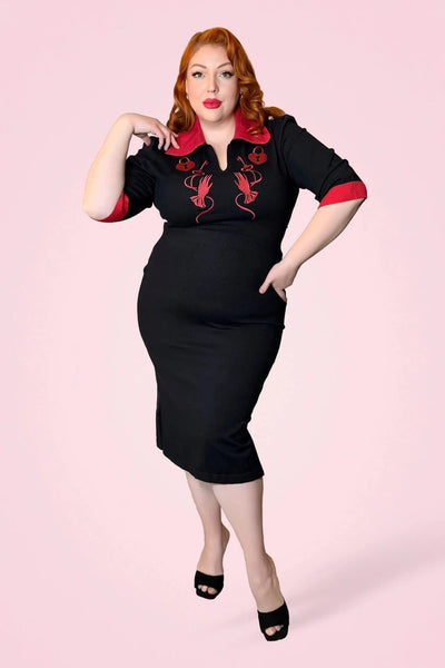 model wearing black sweater knit dress with bright red rolled collar, v-neckline, elbow length sleeves with red cuffs, and embroidered detail across the bodice in matching red of heart-shaped lock, key, and gloved hand. Dress has pencil skirt with side slit and red patent faux leather skinny belt. Shown from front