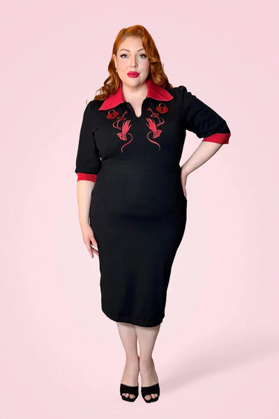 model wearing black sweater knit dress with bright red rolled collar, v-neckline, elbow length sleeves with red cuffs, and embroidered detail across the bodice in matching red of heart-shaped lock, key, and gloved hand. Dress has pencil skirt with side slit and red patent faux leather skinny belt. Shown from front
