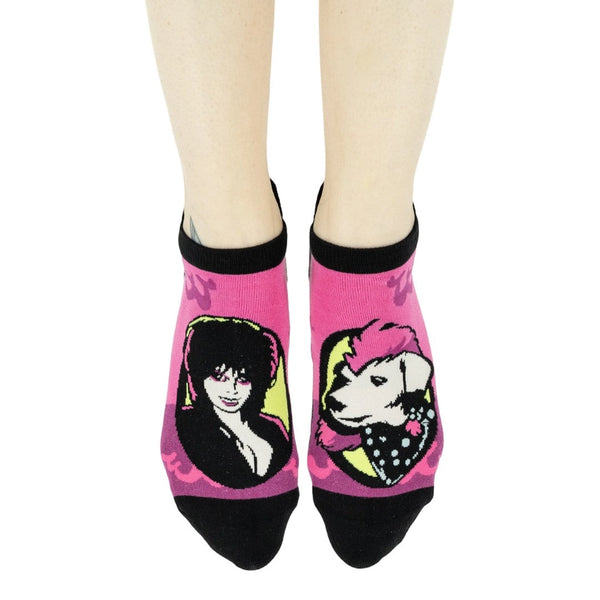 pair of ankle socks in neon pink with black toes, heels, and cuffs and portrait of Elvira in purple sunglasses on one sock and Gonk the dog on the other sock. Shown worn by a model