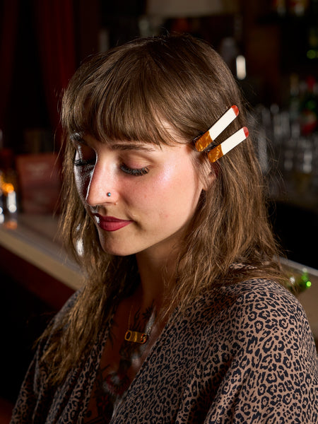 pair of acetate hair clips in the shape of cigarettes with red "lit" ends and marbled brown "filters". Shown worn by model in bar setting