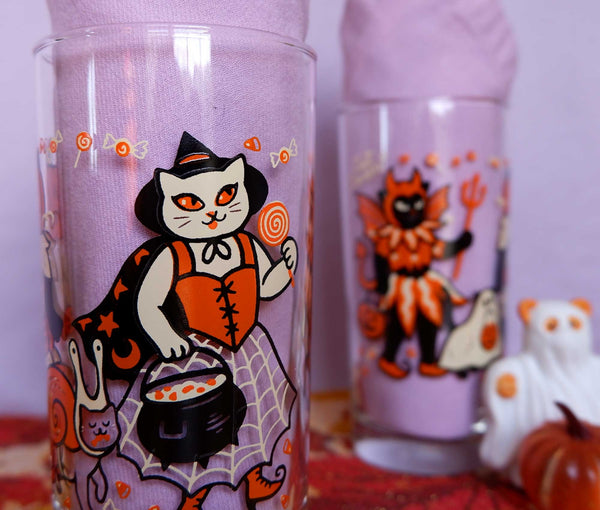 Drinking glasses screen printed with illustrations of cats wearing vintage inspired Halloween costumes. Two shown next to each other on a purple background with one shown in foreground