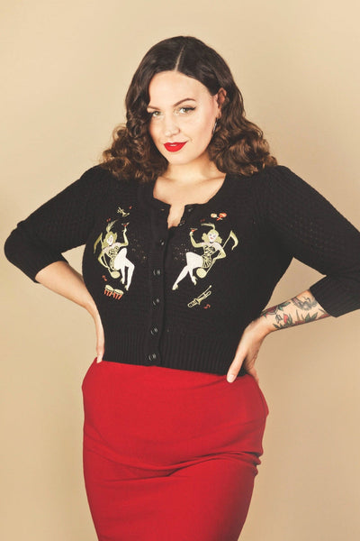 model wearing black cotton pointelle knit cardigan with embroidery detail on both sides of the chest of Vegas style yellow animal print-clad showgirls posed pinup style atop giant golden music notes surrounded by yellow and red musical instruments. Shown from the front 