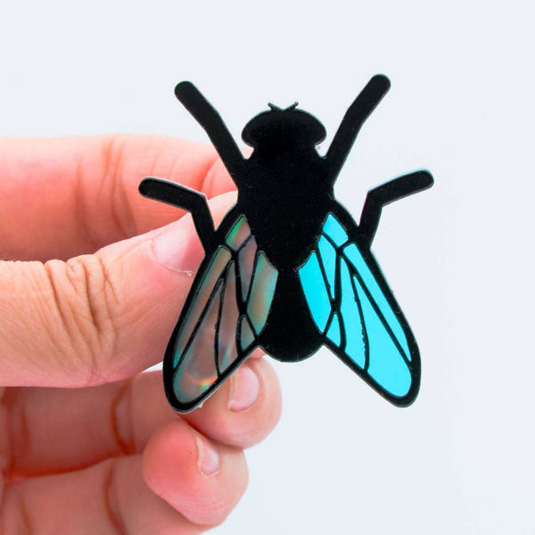 layered acrylic brooch of a black fly with iridescent translucent blue wings. Shown held by hand for scale
