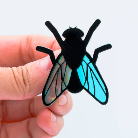 layered acrylic brooch of a black fly with iridescent translucent blue wings. Shown held by hand for scale