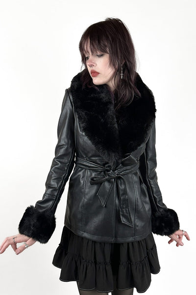model wearing a black faux-leather wrap coat with a large black faux fur shawl collar, matching fur-trimmed cuffs, and a self-tie belt. Shown from front closed