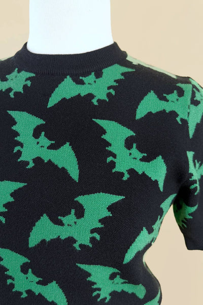 short sleeve acrylic knit sweater with black background and all-over tessellated bright green flying bat print. The top has a crew neck and ribbed hem. Shown from front in close up of pattern