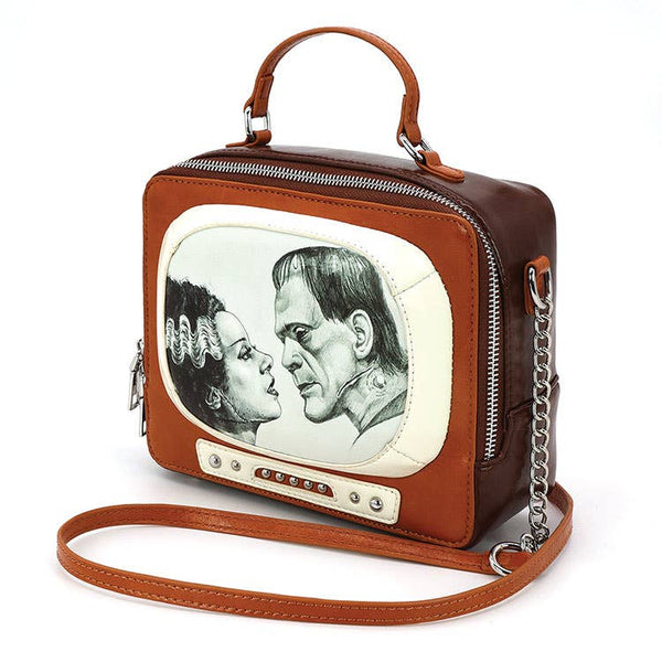 A rectangular novelty purse in the shape of a vintage television set in light brown and cream faux leather with silver details. Has a “screen” with a black and white glow-in-the-dark image of Frankenstein and his Bride and faux knob controls. Has a brown handle and detachable matching cross body strap. Shown from the side