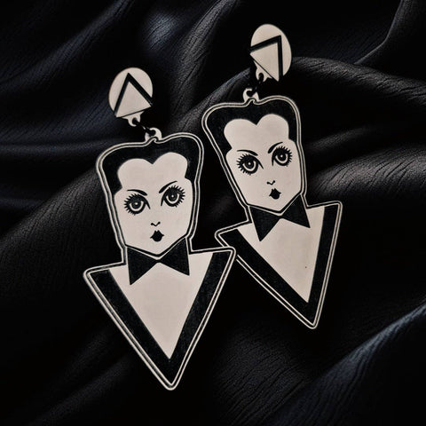 pair of black and white laser-cut acrylic drop earrings in the shape of a Kewpie Doll-style Klaus Nomi wearing their geometric black and white tuxedo outfit. Connected to round white charms with a black and white triangle. Shown on black cloth