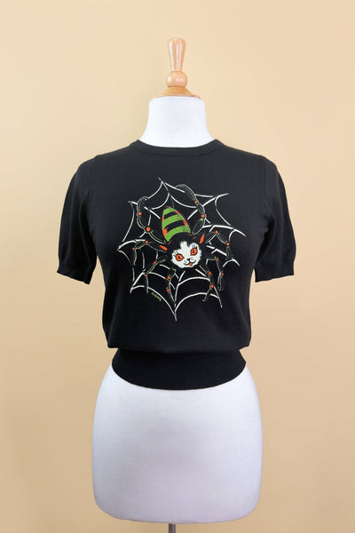 black crew neck short sleeved sweater with white, bright orange, green, and black printed image of cat with spider body and legs smiling on top of a spider web. Shown on dress form