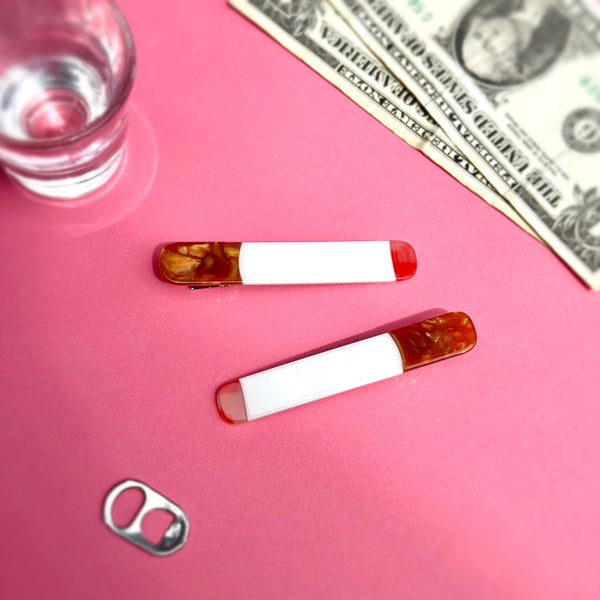 pair of acetate hair clips in the shape of cigarettes with red "lit" ends and marbled brown "filters". Shown flat