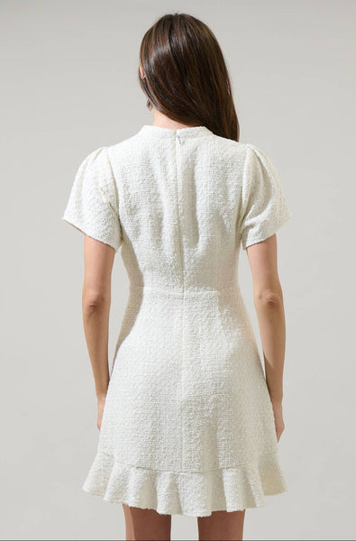 model wearing white textured tweed woven mini dress with mock neck, short sleeves with slightly puffed shoulders, bodice with vertical ruffled bib style and three black bows running down the middle. Dress has a-line skirt ending above the knee in a wide gathered ruffle. Shown from the back