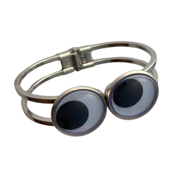 Silver metal hinged bracelet with large two black and white plastic googly eye adornments on front. Shown flat