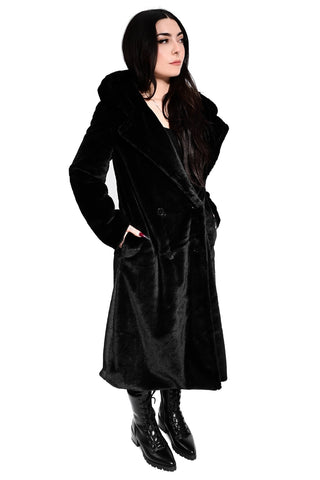 model wearing a black faux fur coat with wide lapel and oversized faux fur lined hood. Coat has double black plastic button closure and side seam pockets. Shown from three quarter angle with hands in pockets
