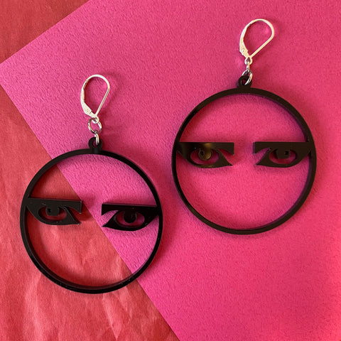 Laser-cut black acrylic dangle earrings in the shape of 2” thin hoops with illustration of Siouxsie Sioux’s eyes horizontally across each hoop. Attaches to ear with sterling silver lever back style hooks. Shown flat on a neon pink background