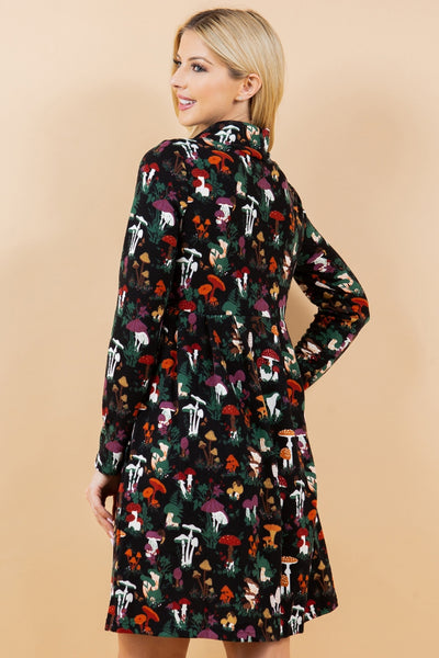 model wearing a knit long sleeved dress in a black background multicolored woodsy mushroom print. it has a cowl style neck, pockets, and knee length skirt. Shown from the back