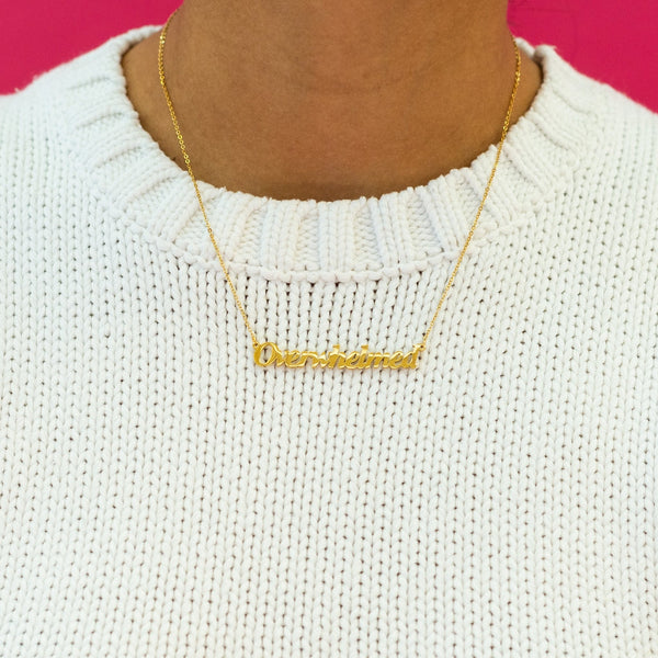 Gold covered stainless steel link chain necklace with 'Overwhelmed' nameplate pendant shown worn by model wearing white crewneck sweater