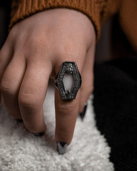 A model wearing a large ring with a hexagonal top decorated with filigree details and a large white semi translucent middle. Seen from the front