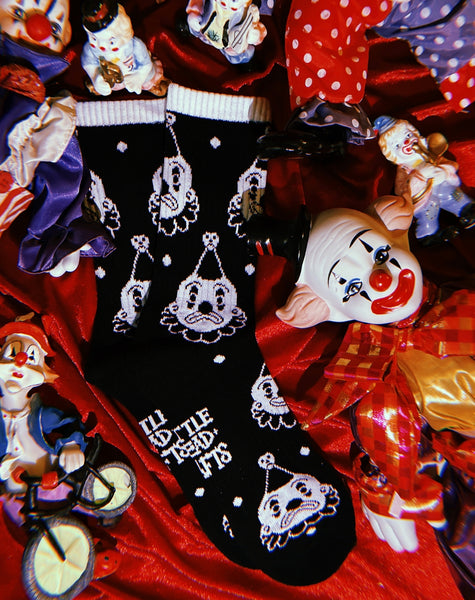 black cotton crew socks with white cuffs and an all over pattern of a white and black clown with either a smiling or frowning face. Shown flat on background of ceramic clown statues