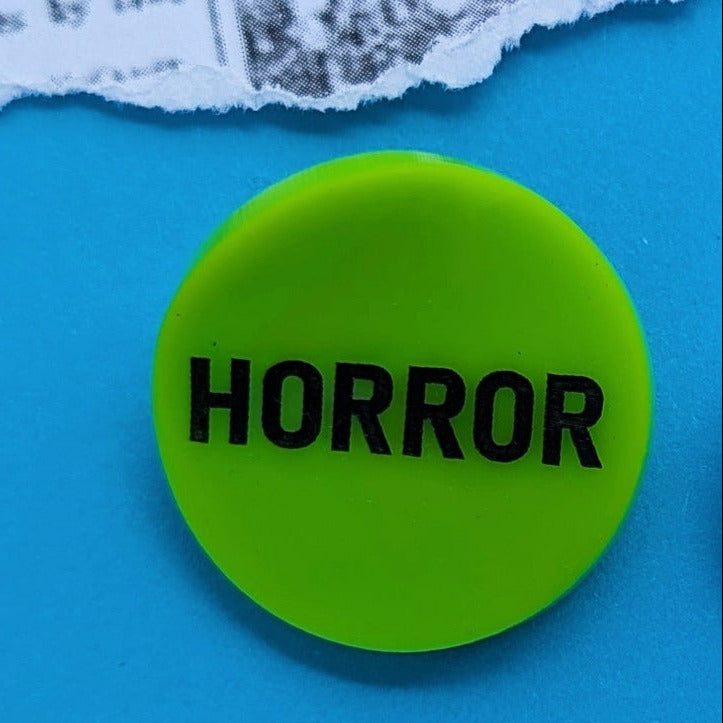 round neon green acrylic brooch with slightly concave shape and "HORROR" in black shown on neon blue torn newspaper background
