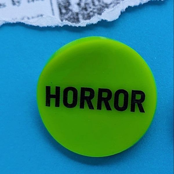 round neon green acrylic brooch with slightly concave shape and "HORROR" in black shown on neon blue torn newspaper background