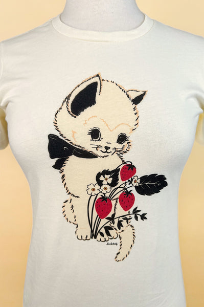 Fitted off-white t-shirt with printed image of a cream-colored kitten with black bow around its neck alongside a blossoming strawberry plant with three berries. Shown on a dress form from the front in close up