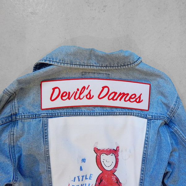 Back of a light blue denim jacket featuring a white rectangular patch with red embroidered text reading 'Devil's Dames' 