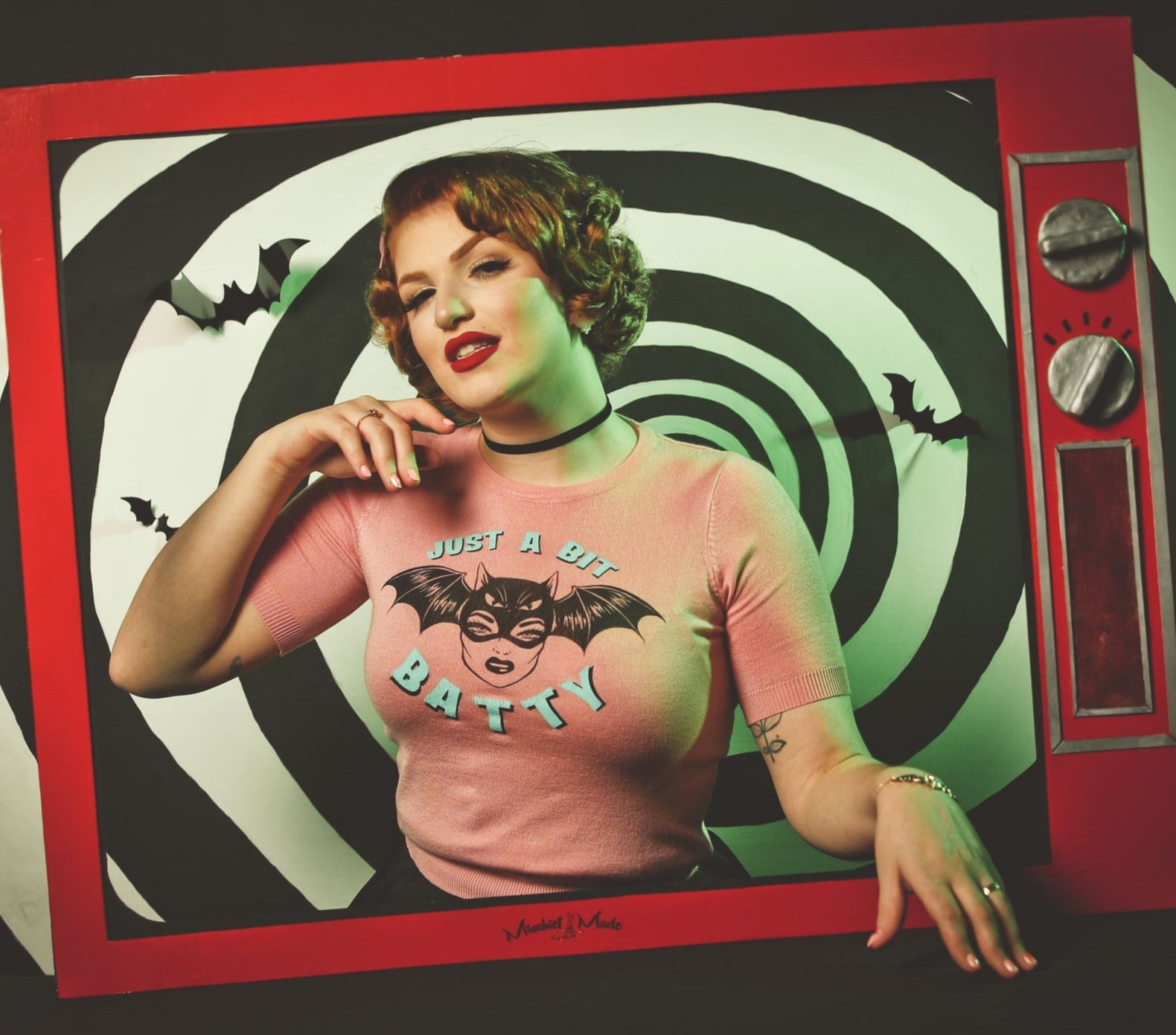Model standing in a cardboard set meant to look like an old fashioned television screen. Wearing pink crew neck short sleeved sweater with retro woman wearing a bat mask and “Just a Bit Batty” written in bright blue