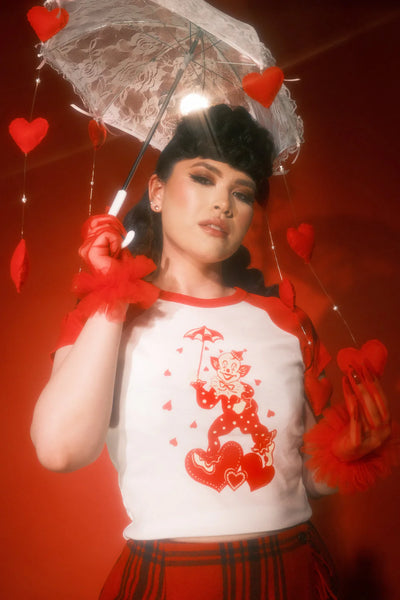Short sleeved crop white t-shirt with red raglan sleeves and red & cream illustration of a clown wearing a polka dotted outfit holding an umbrella standing on a trio of hearts and surrounded by hearts raining down. Shown on a model wearing a plaid skirt and holding a white lace parasol.