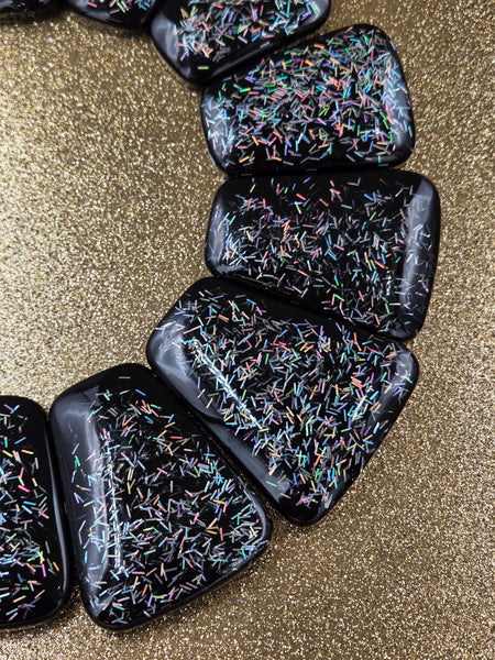 close up of bib necklace made of segments of flat black resin rounded trapezoid shapes with iridescent bar glitter