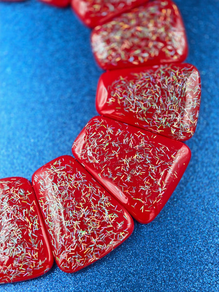close up of bib necklace made of segments of flat red resin rounded trapezoid shapes with iridescent bar glitter