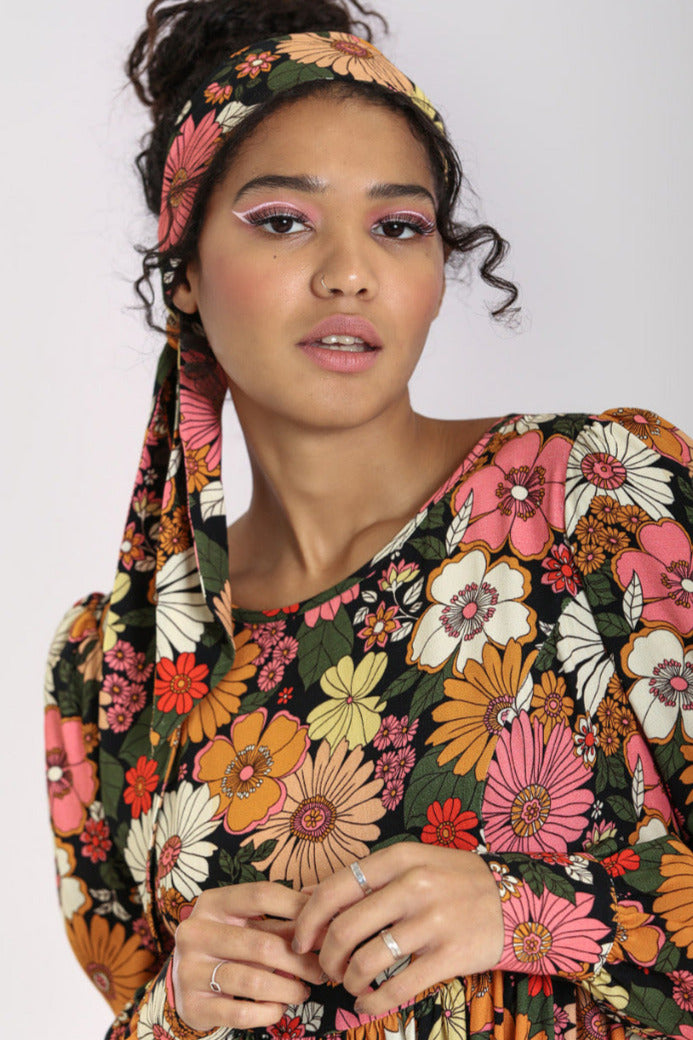 Model wearing long rectangular hair scarf in textured crepe fabric with black background and retro 60’s/70’’s floral pattern in shades of orange, pink, green, yellow, and white