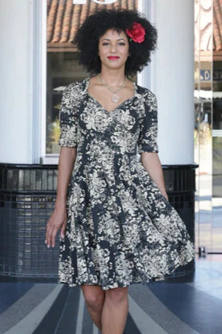 black stretch cotton fit and flare dress in an allover printed damask style floral pattern in cream featuring a surplice bodice with Queen Anne neckline, puffed shoulder short sleeves, and full knee length skirt. shown worn by a model