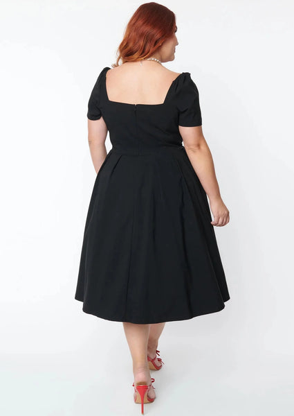 black stretch bengaline dress featuring short sleeves, an open sweetheart neckline, decorative button detail at the bust, fitted bodice, and pleated full just below the knee length skirt. shown back view on a model.
