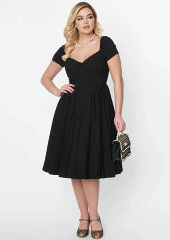 black stretch bengaline dress featuring short sleeves, an open sweetheart neckline, decorative button detail at the bust, fitted bodice, and pleated full just below the knee length skirt. shown on a model.