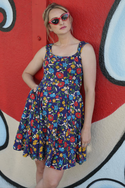 Model wearing cotton knit stretch dress in a dark purple-y blue background sketch style floral allover pattern with red, blue, orange, yellow, and off-white wildflowers. Dress has rounded square neckline, fitted bodice, full flared just-above-the-knee skirt and side seam pockets