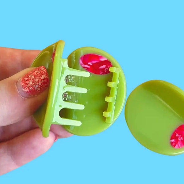 Two light green acetate mini hair clips in the shape of an olive with red marbled detail as a “pimento” middle, one held open in a hand. Shown on light blue background