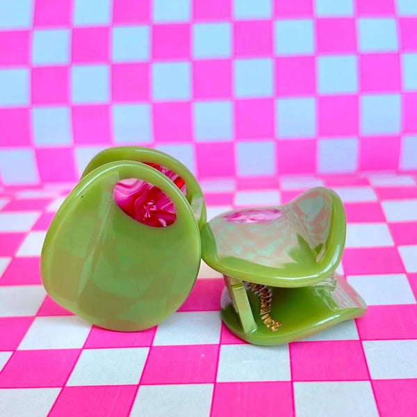 Two light green acetate mini hair clips in the shape of an olive with red marbled detail as a “pimento” middle. Shown on a pink checker print background