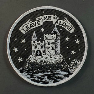Round embroidered patch picturing a castle illustrated in white surrounded by a moat with the caption “LEAVE ME ALONE” in white ribbon