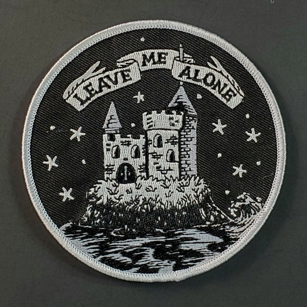Round embroidered patch picturing a castle illustrated in white surrounded by a moat with the caption “LEAVE ME ALONE” in white ribbon