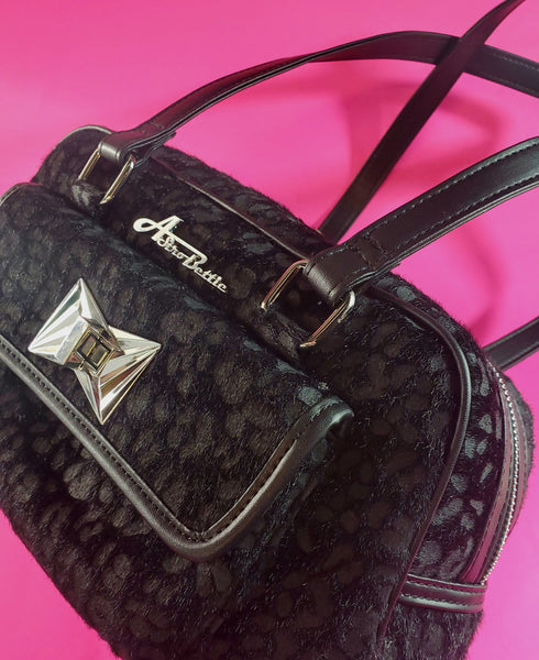 Trapezoid-shaped purse in a black on black leopard pattern printed on fuzzy faux pony with matching matte faux leather trim and shoulder straps. The large exterior pocket on the front features an Art Deco inspired silver metal clasp and the Astro Bettie logo in silver metal