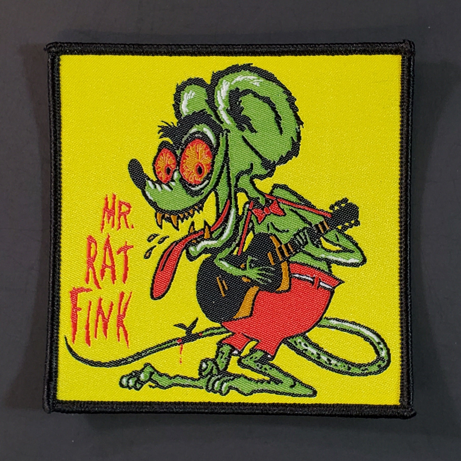 A green, red, white, orange, and black woven patch with a black embroidered border with an illustration of Rat Fink playing an acoustic guitar with the caption “MR. RAT FINK” in red lettering beside