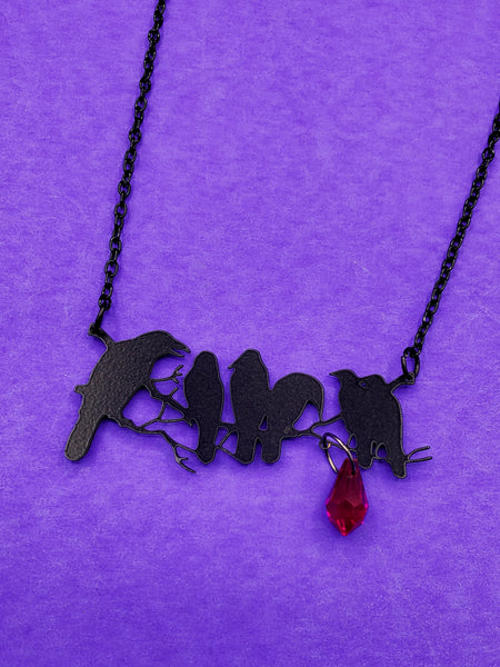 necklace with large black enameled metal pendant of six crows seen in silhouette on a branch with attached faceted red teardrop bead. On a 17" black enameled link chain