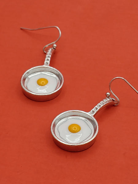 Pair of dangle earrings, each in the shape of a silver metal frying pan with white and yellow enamel sunny side up egg inside