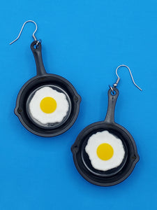 pair of dangle earrings with charms in the shape of cast iron frying pans with sunny side up eggs in each pan